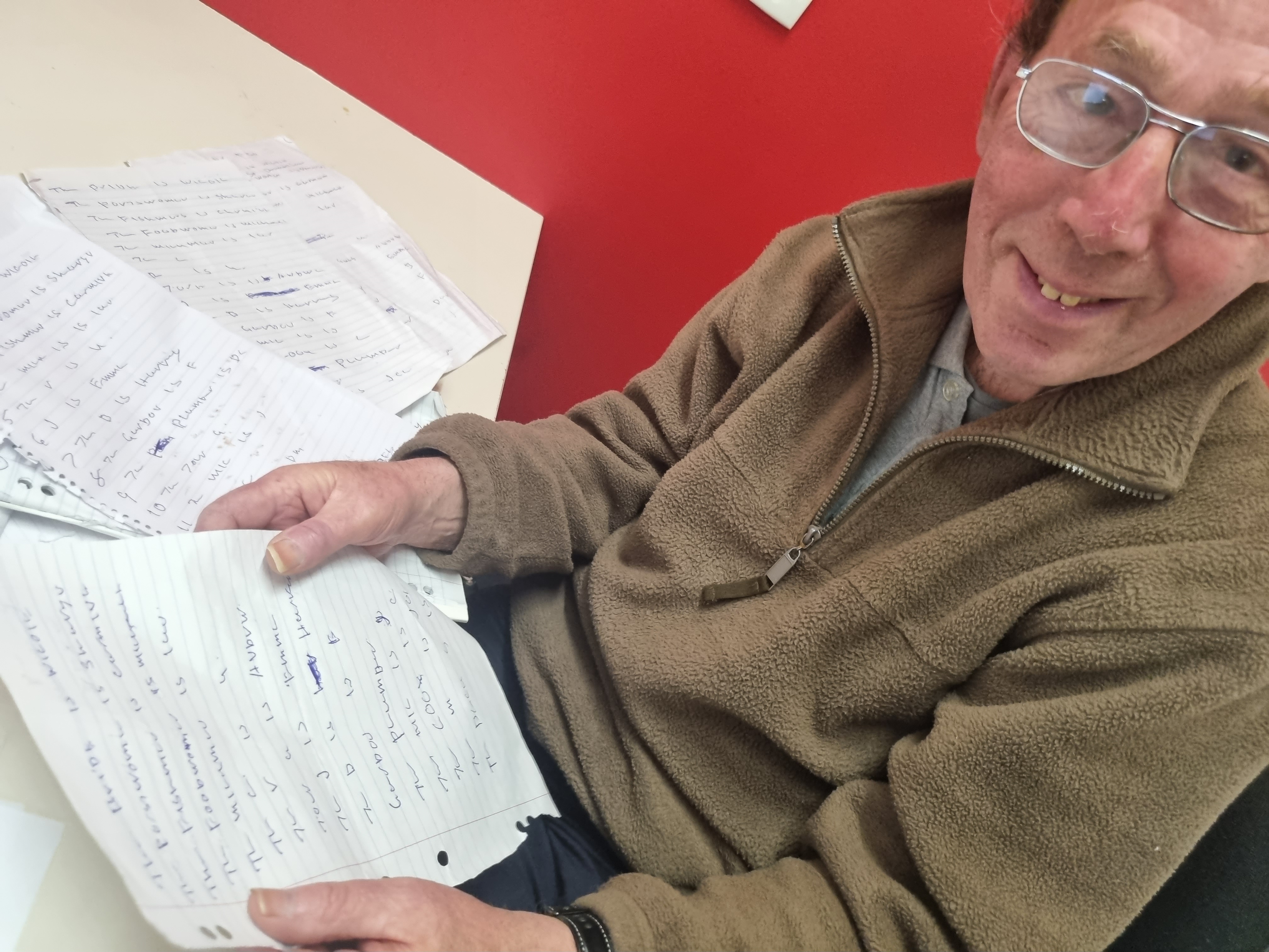 A portrait of Bill holding his original hand written script in hand, looking up to camera with a big smile. He wears glasses and is sitting a desk in a meeting room that has red walls.