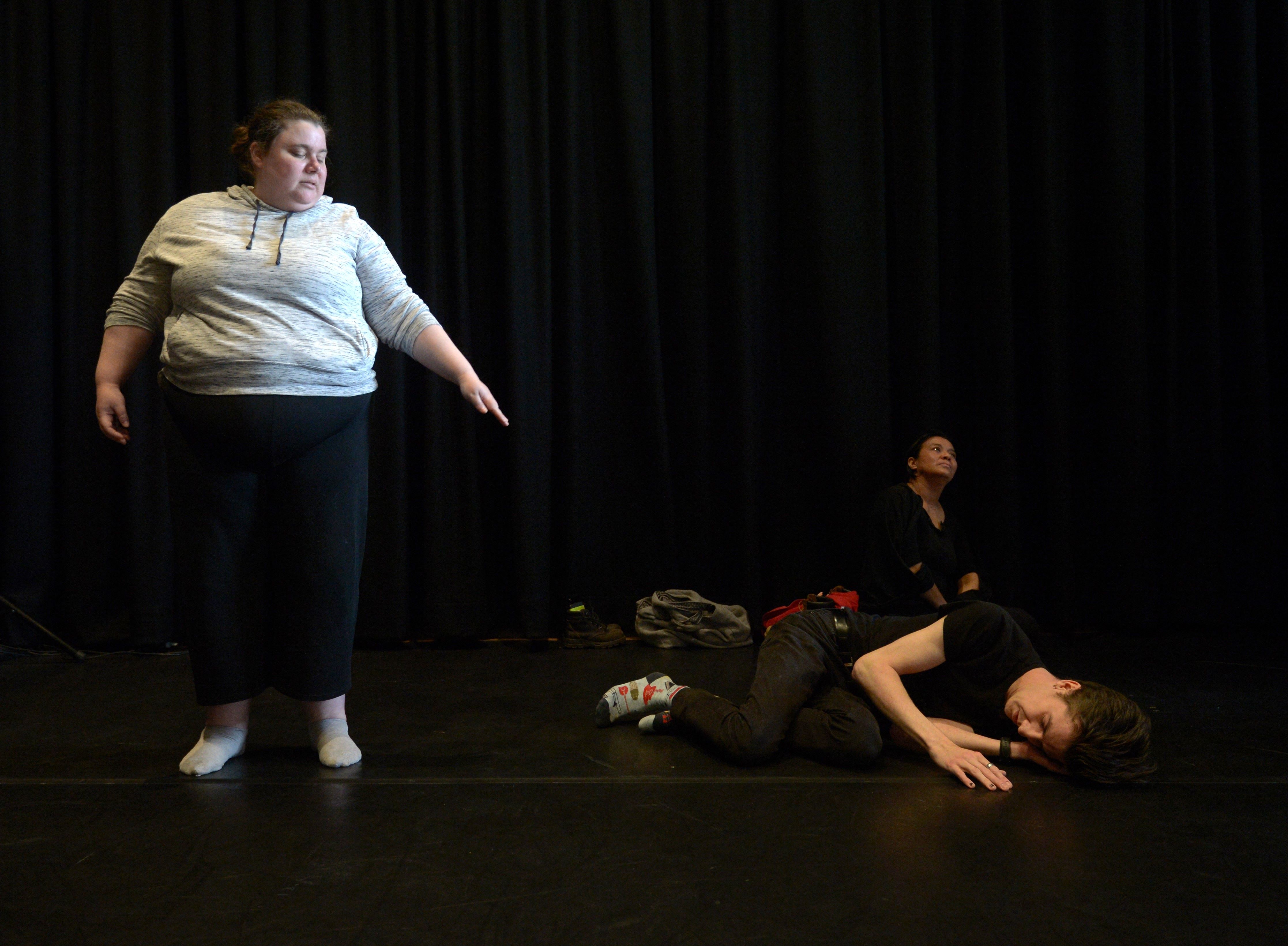 An image of Alex Craig, lit in natural light, and Jeremy Lowrencev on stage as part of Arts House and VV's CultureLab project called Disrupting sighted ableism. In background seated against black curtain is other co-performer Imogen Yang. Alex stands to the left of Jeremy who is curled on floor. Alex's left arm is extended out from their side in diagonal line to his feet. Both performers eyes are closed. Photo by Carla Gottgens.