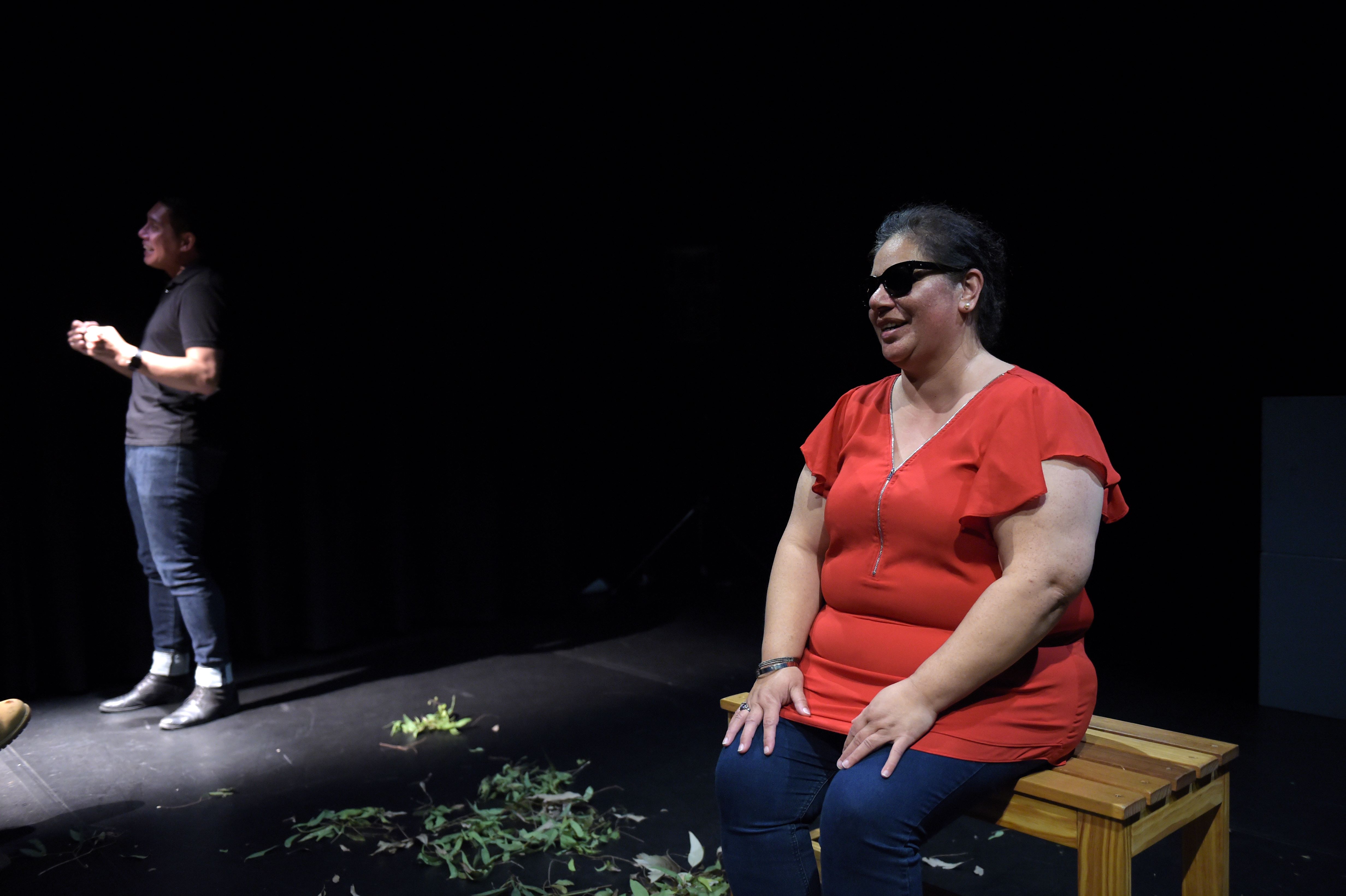 Performer Vanessa Nanai on stage seated at the wooden bench she made, smiling, wearing fire engine red top, blue jeans, and sunglasses, both palms resting on her knees. To her left standing is Auslan interpreter Marc Ethan in bright light signing, and on floor eucalyptus are strewn. From Arts House and VV's CultureLab project Disrupting sighted ableism. Photo by Carla Gottgens.