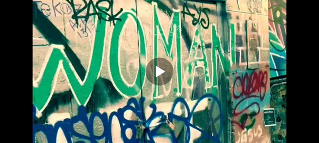 Screenshot in landscape orientation from Sammi’s exploratory ‘theme-of-green’ short video of the CBD Melbourne of a wall featuring layered and graffiti’ed tags and designs. The main focus of this shot is the word WOMAN in large capital letters sprayed with green paint, with a thin white outline. The triangular shaped play icon function is in the centre of the image.  
