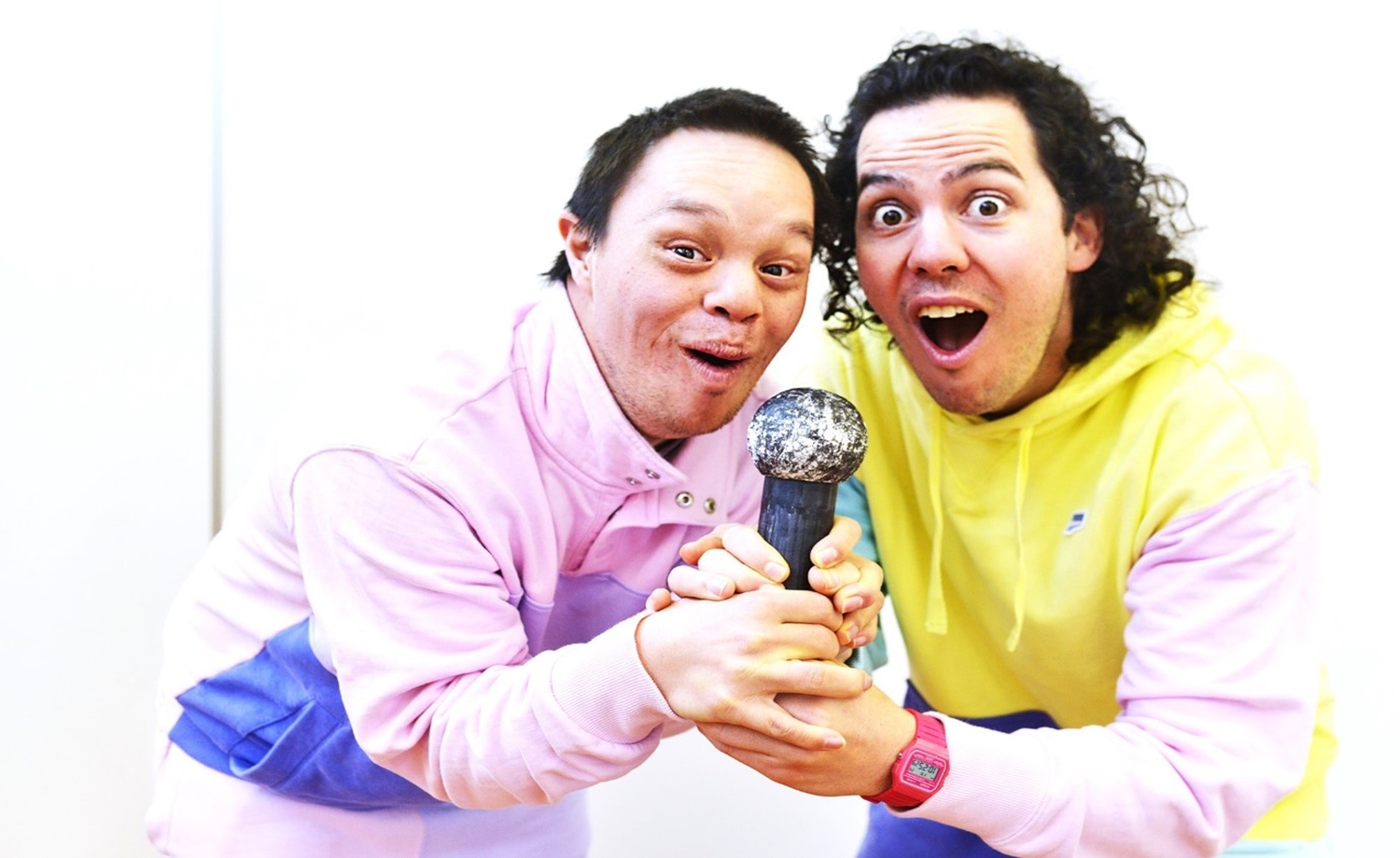 Two male performers in pastel coloured tracksuits side by side with both sets of hands clutching at one makeshift microphone. Both have friendly eyes smiles beckoning you to come to their new show. Image from Breakaway by William Bailey and David Maney, as part of VV's mentoring programs. Photo David de Roach 2022