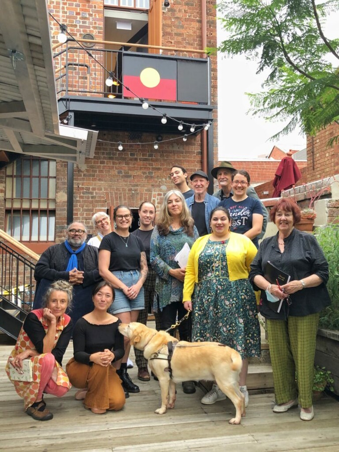 With permission La Mama Theatre, image by Georgina Capper, thirteen people and an assistance dog stand and crouch in a group on decking in front of the rebuilt two storey La Mama HQ theatre. The Aboriginal flag is attached to the upper floor railing, a string of lights is suspended above their heads, and a leafy branch of a jacaranda tree stretches over them. They are all smiling. From VV's audio description and tactile tour training sessions for staff, co-delivered with Olivia Muscat.